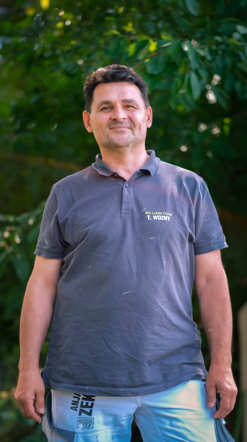 Man standing outside wearing a dark polo shirt and light pants with green foliage in the background.