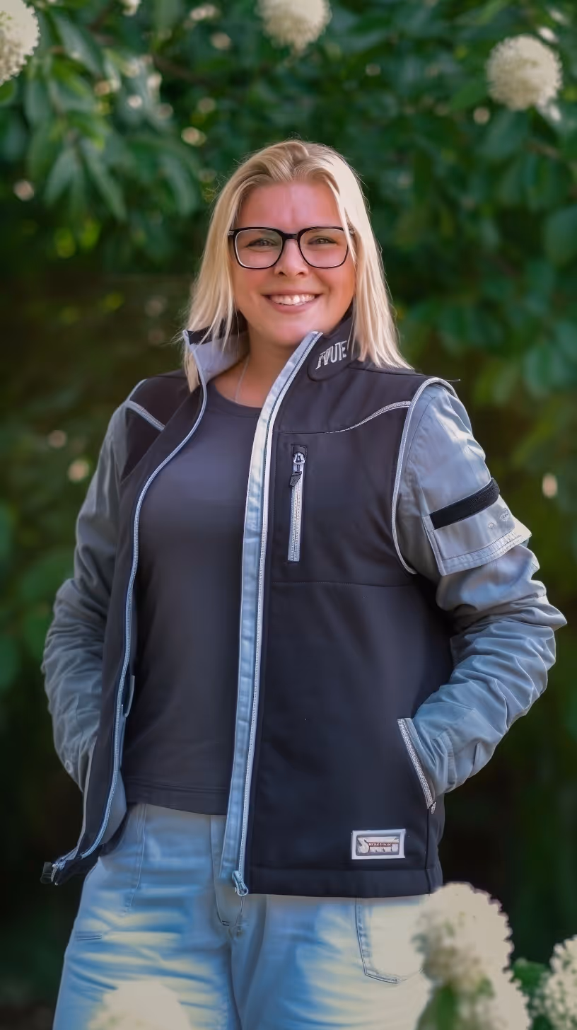 Smiling blonde woman wearing glasses and a black vest standing outdoors with green foliage behind.