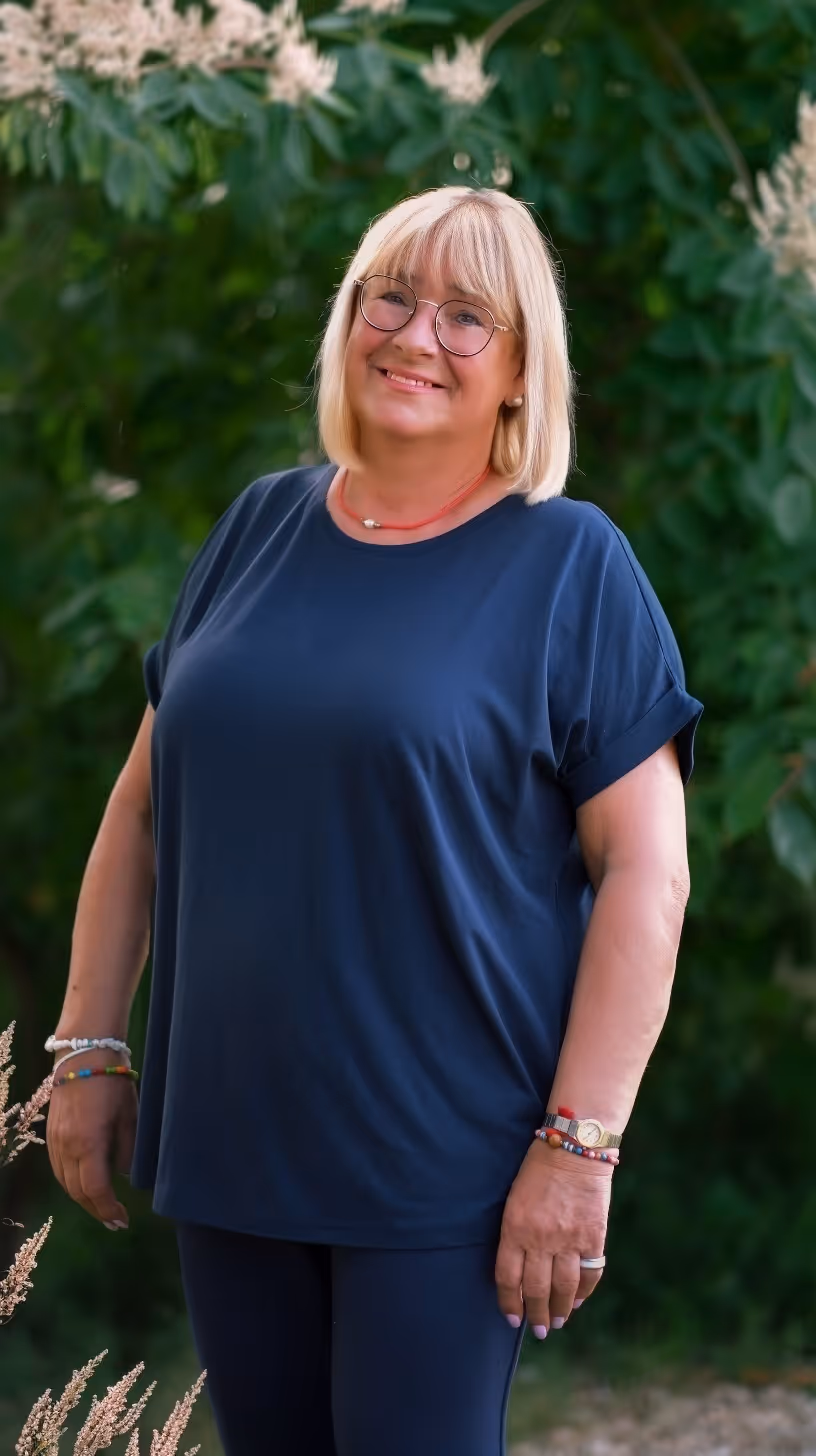 Smiling blonde woman wearing glasses and a navy shirt stands outside with green foliage in background.