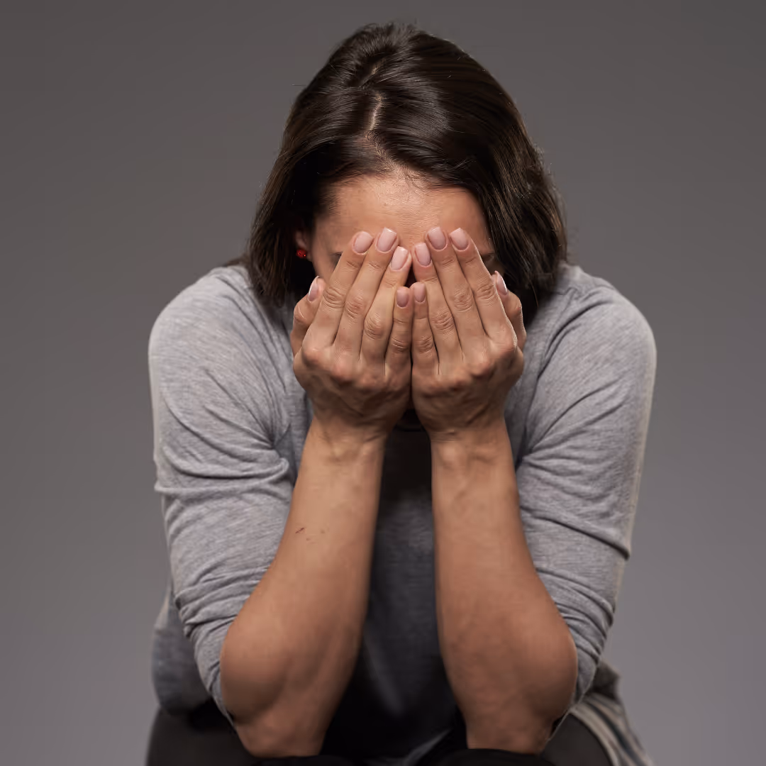 Frau in grauem Pullover hält sich die Hände vors Gesicht - Ausdruck von Stress oder emotionaler Belastung