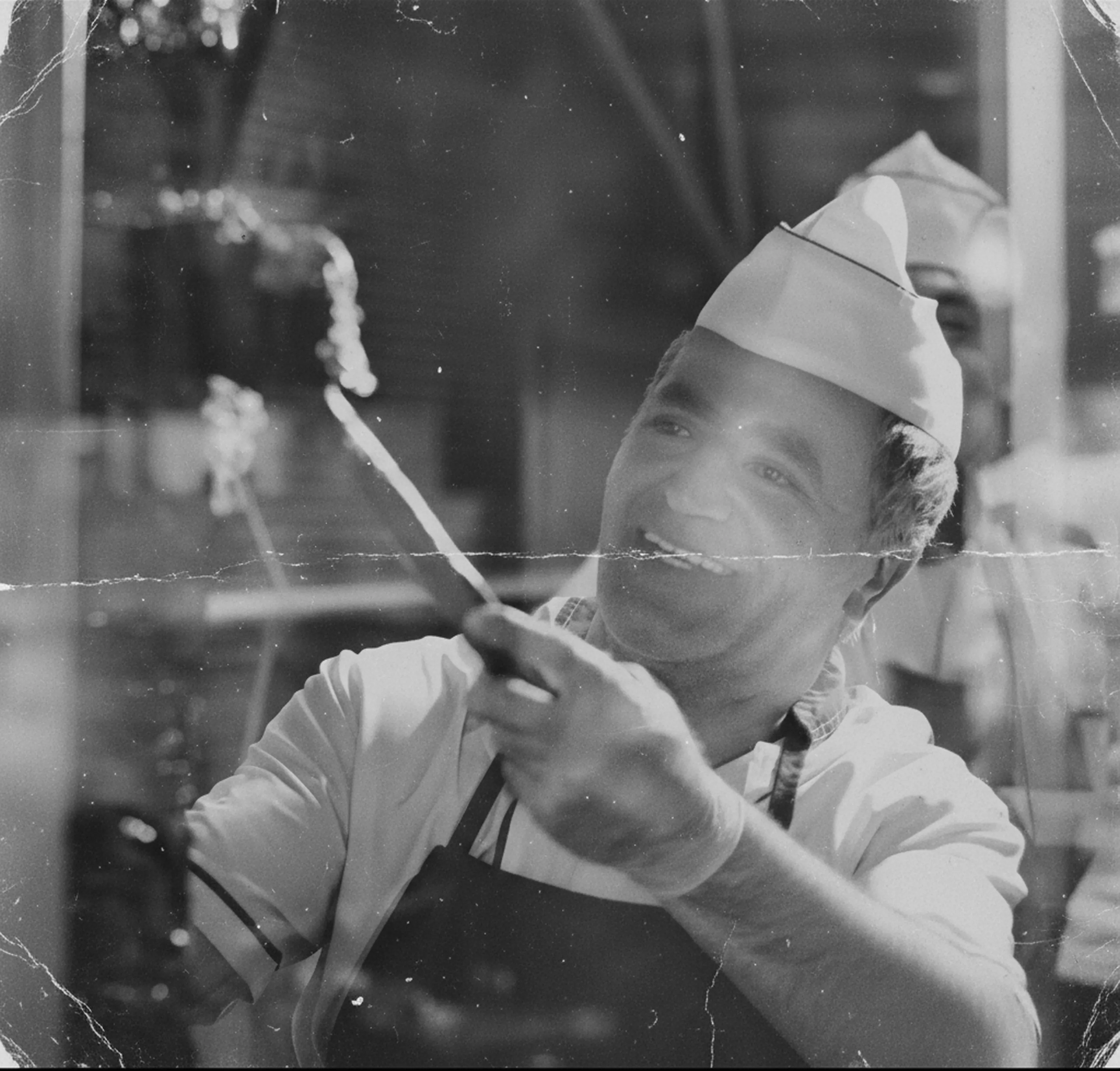 A black and white photograph with a blurry background (bokeh) of a smiling craftsman or cook (wearing a white hat and apron).