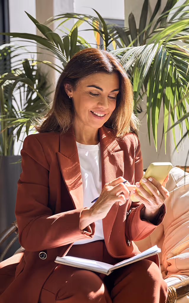 Woman in brown suit sitting in a cushioned wicker chair using a smartphone with a notebook on her lap, surrounded by indoor plants.