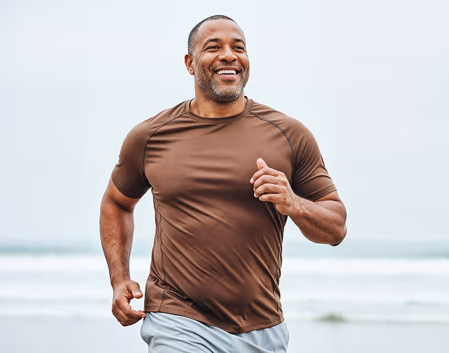 Smiling man wearing brown shirt jogging outdoors near a body of water.