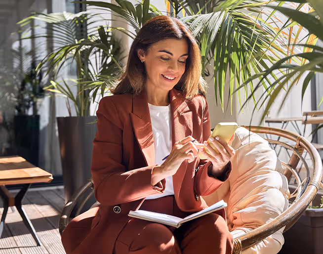 Woman in brown suit sitting in a cushioned wicker chair using a smartphone with a notebook on her lap, surrounded by indoor plants.