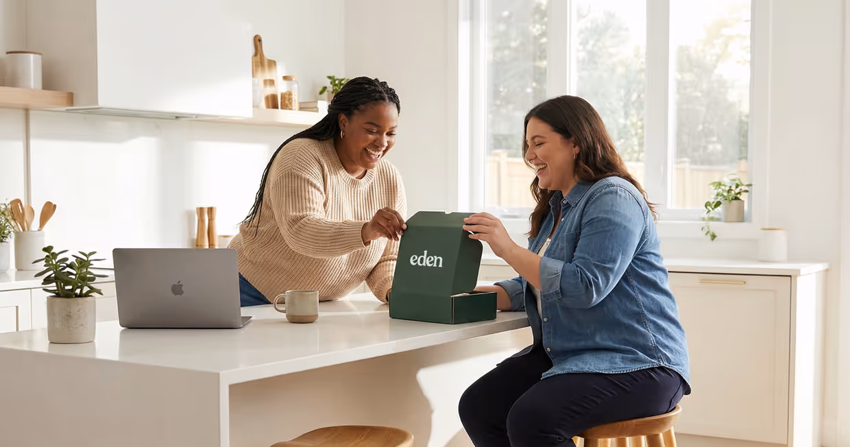 Two women smiling and opening a green box labeled Eden in a bright kitchen with a laptop on the counter.
