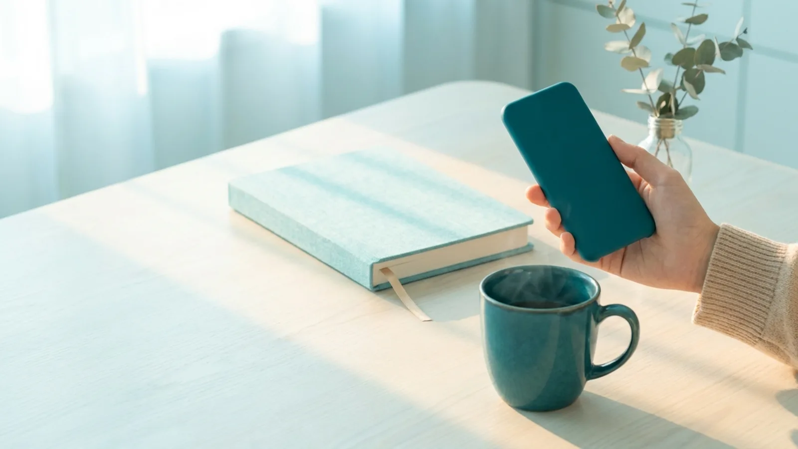A hand holding a phone above a mug next to a paper planner in soft morning light