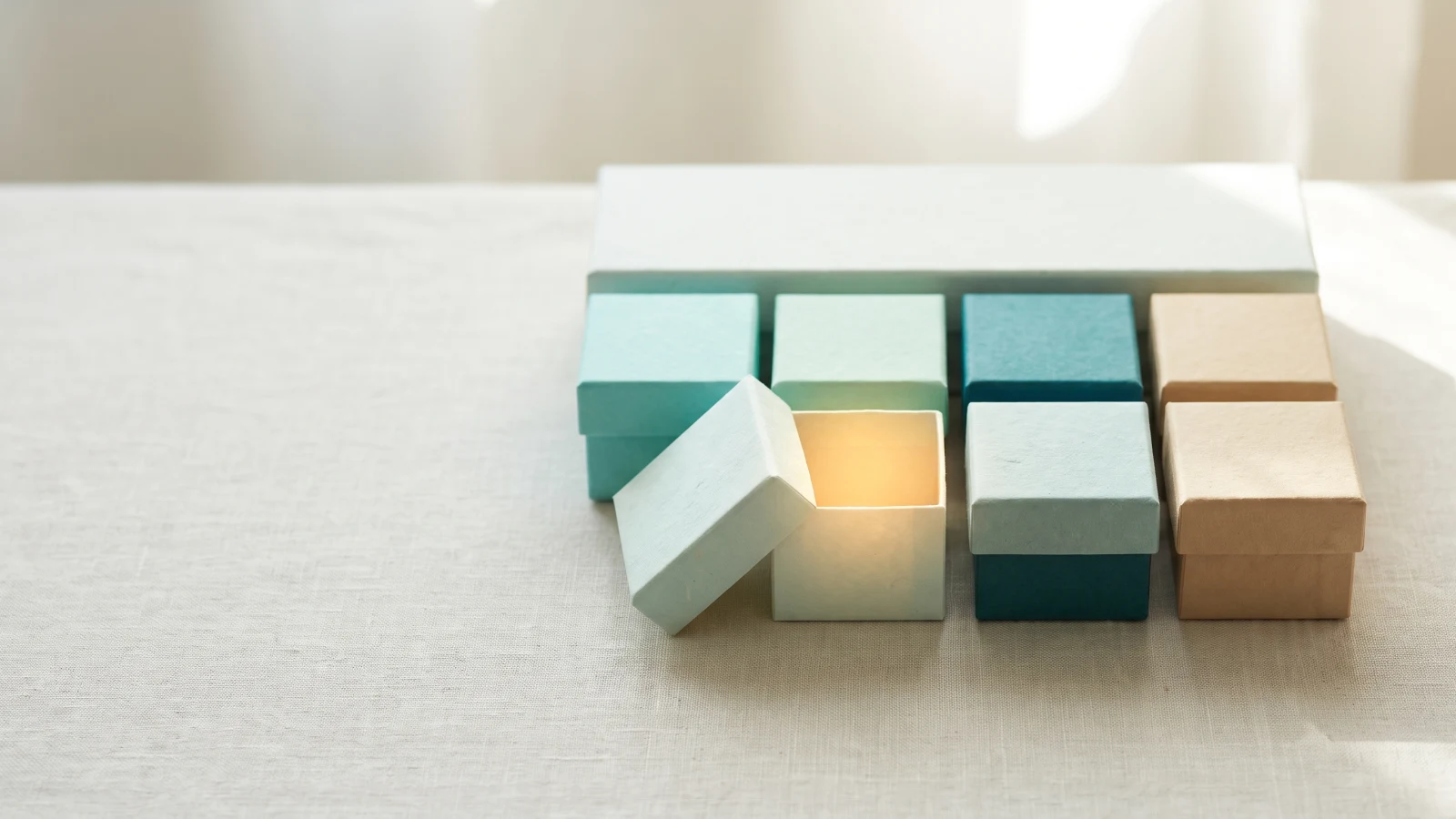 Eight small paper boxes in teal, mint and sand under one long box, a single lid lifted with warm light inside