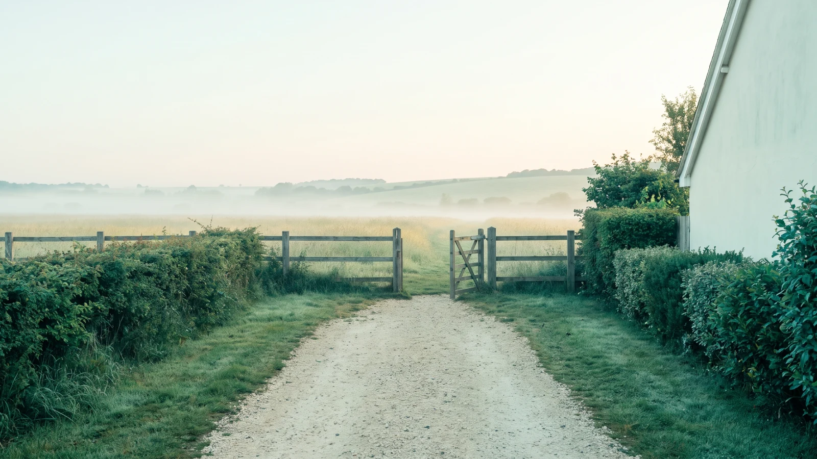 Gravel lane between hedges ending at an open wooden gate onto a misty meadow at dawn, a white cottage wall beside it