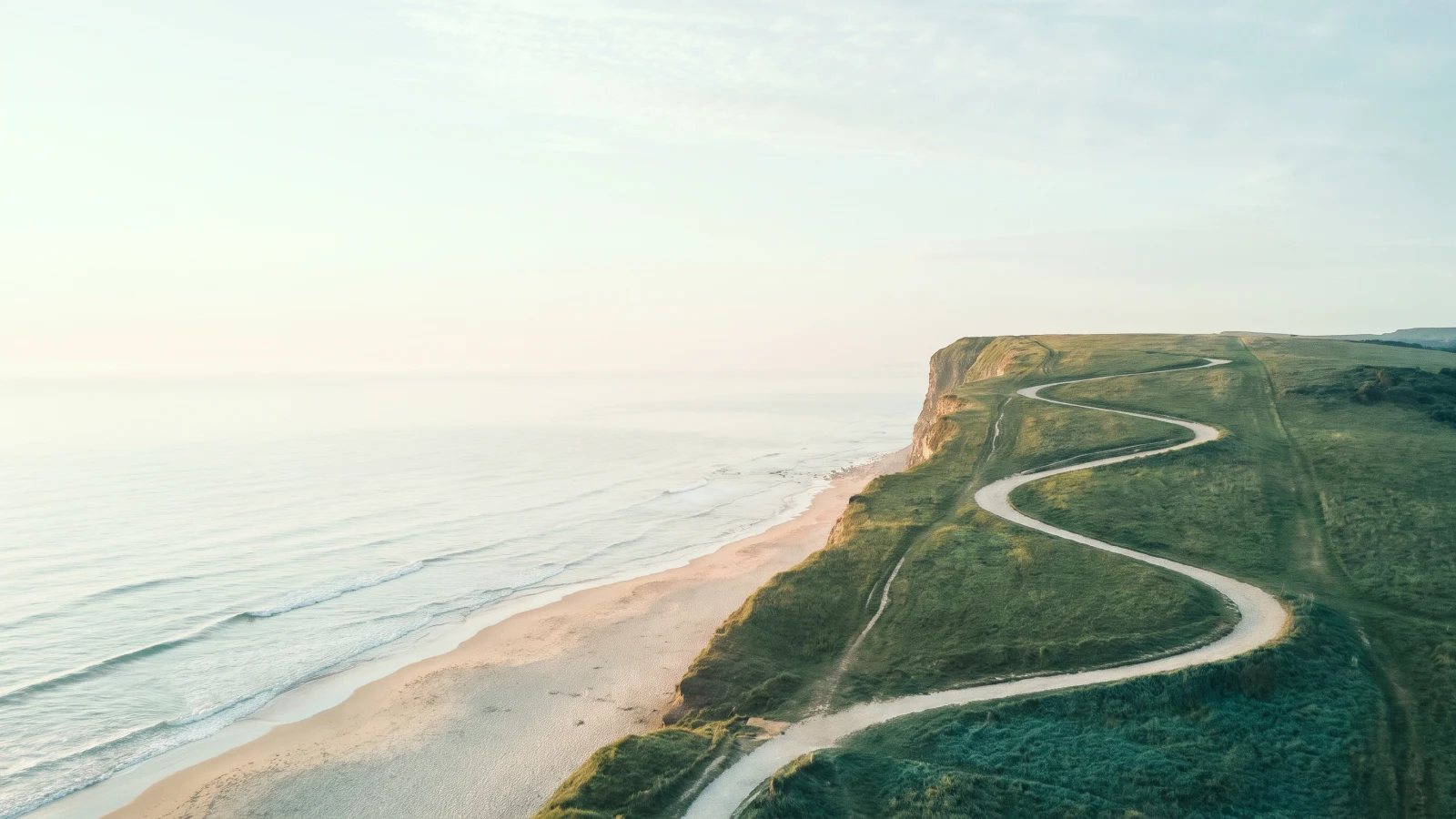 Winding footpath in gentle switchbacks across a green coastal headland beside a cliff edge above a calm beach