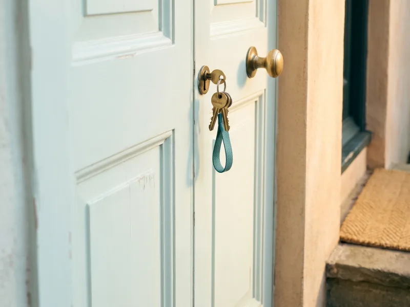 Bunch of brass house keys on a teal strap left hanging in the lock of a pale mint front door in warm morning light