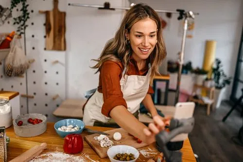 Smiling woman in an apron cooking and filming with a smartphone on a tripod in a kitchen.