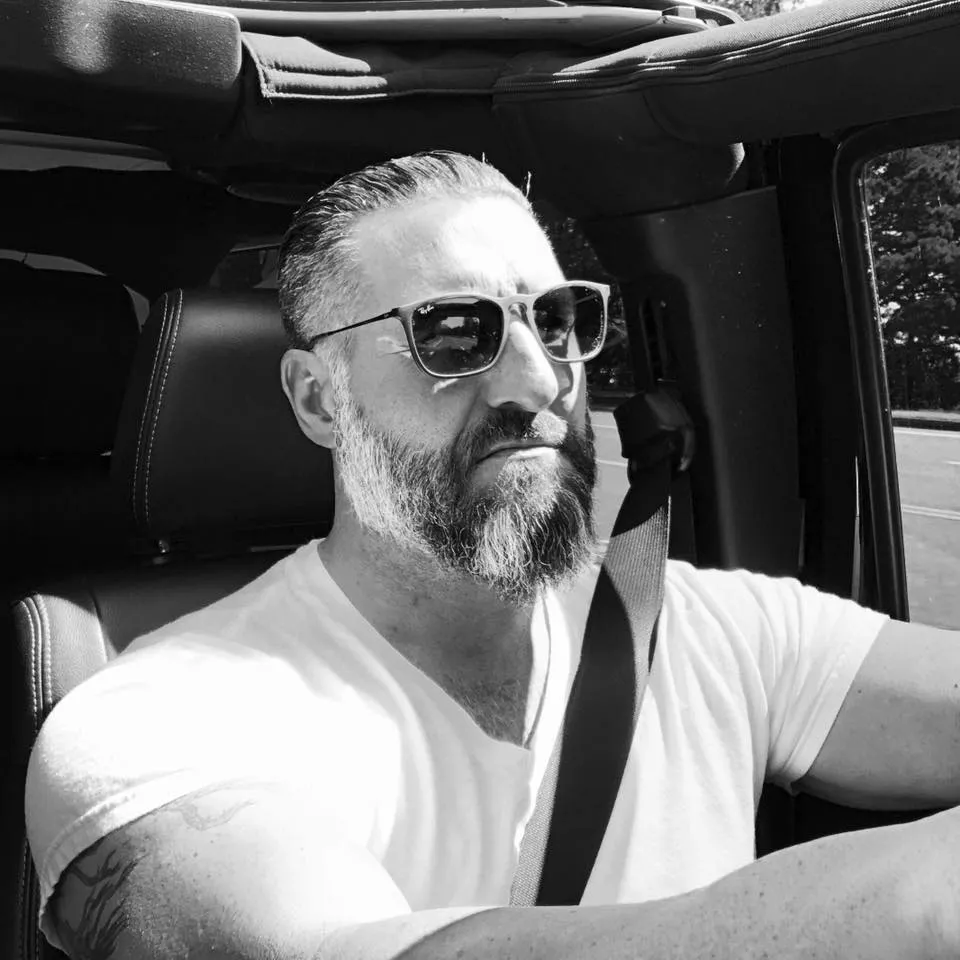 Bearded man wearing sunglasses and a white t-shirt driving a car with a seatbelt fastened.