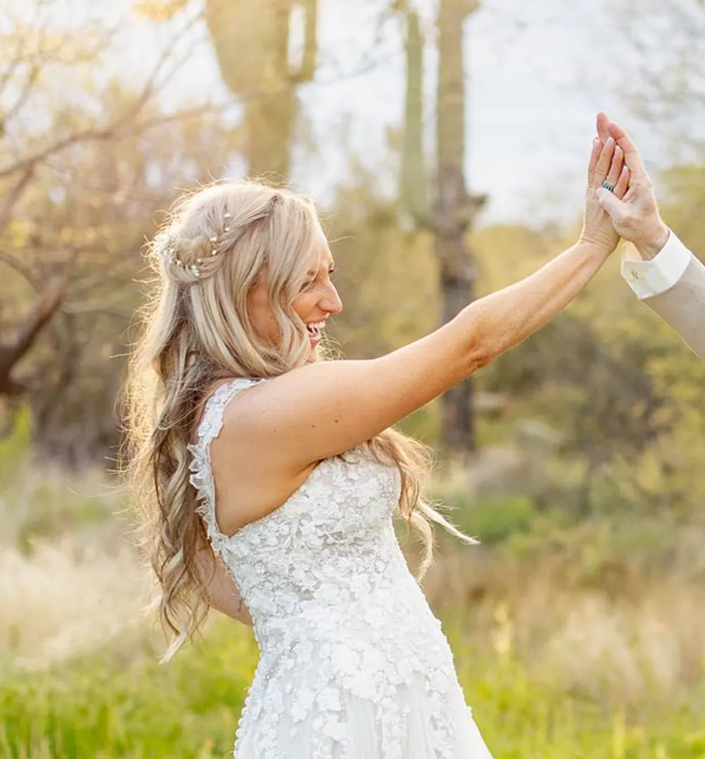 Bride in a white lace wedding dress smiling and giving a high-five outdoors in a sunlit natural setting.