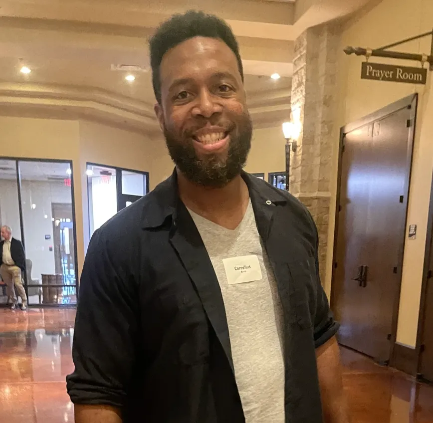 Smiling man with a beard wearing a dark jacket and gray shirt indoors near a sign that says Prayer Room.