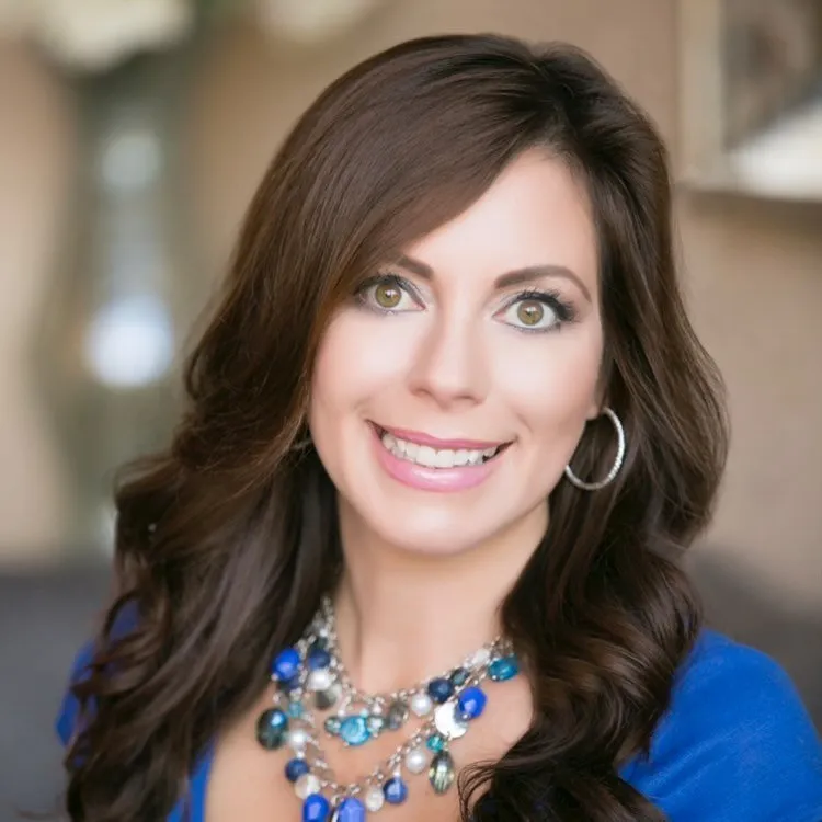Smiling woman with long brown hair wearing a blue top and a multi-colored beaded necklace.