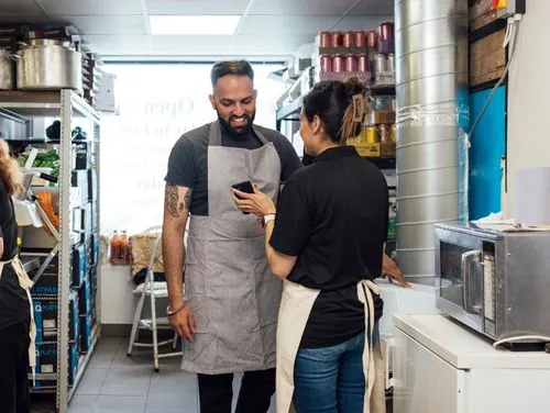 Two restaurant workers in aprons smiling and looking at a phone in a kitchen storage area.