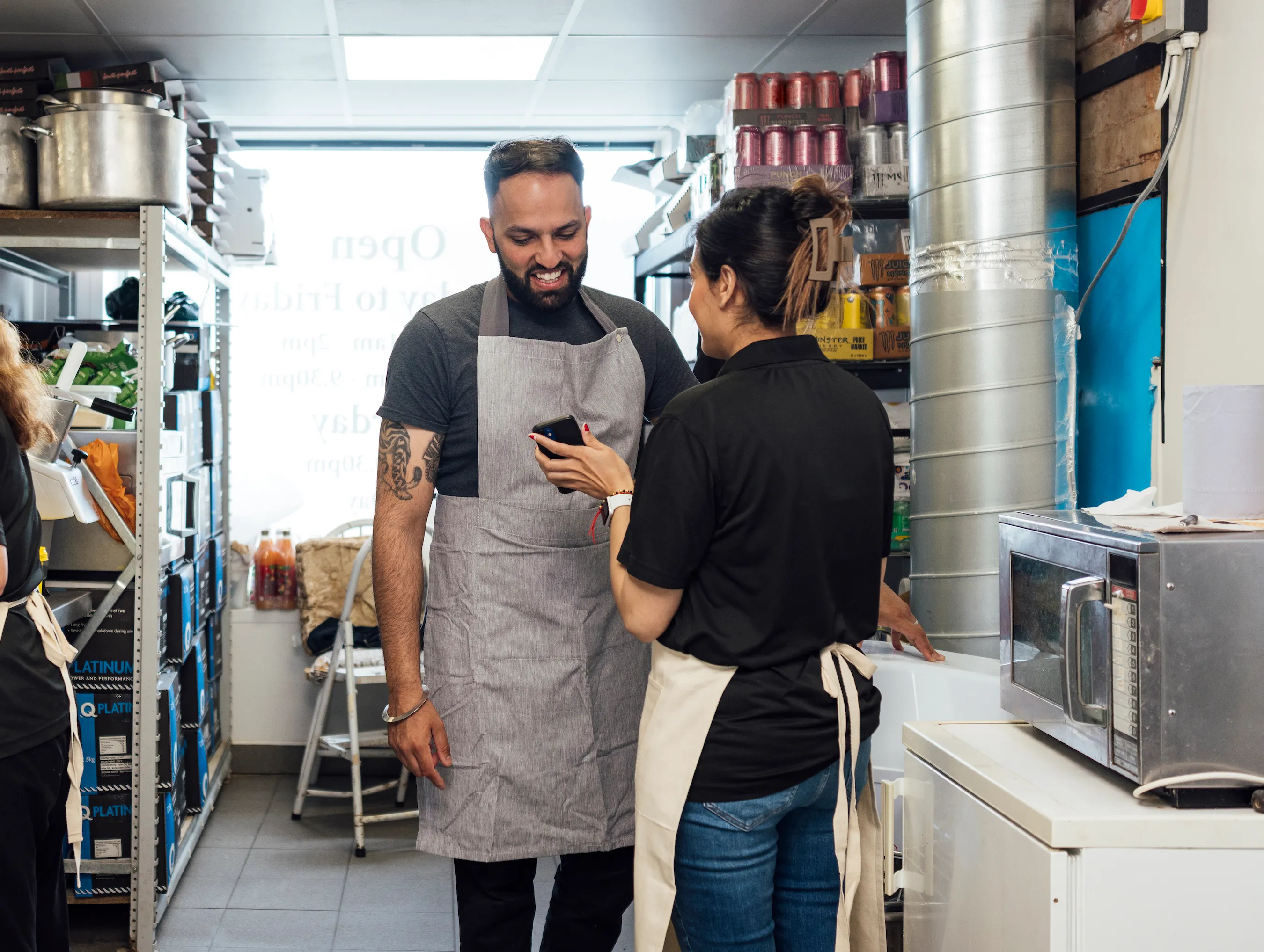 Two restaurant workers in aprons smiling and looking at a phone in a kitchen storage area.