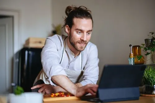 Man in a kitchen wearing an apron, smiling and using a tablet on a wooden table with chopped vegetables.