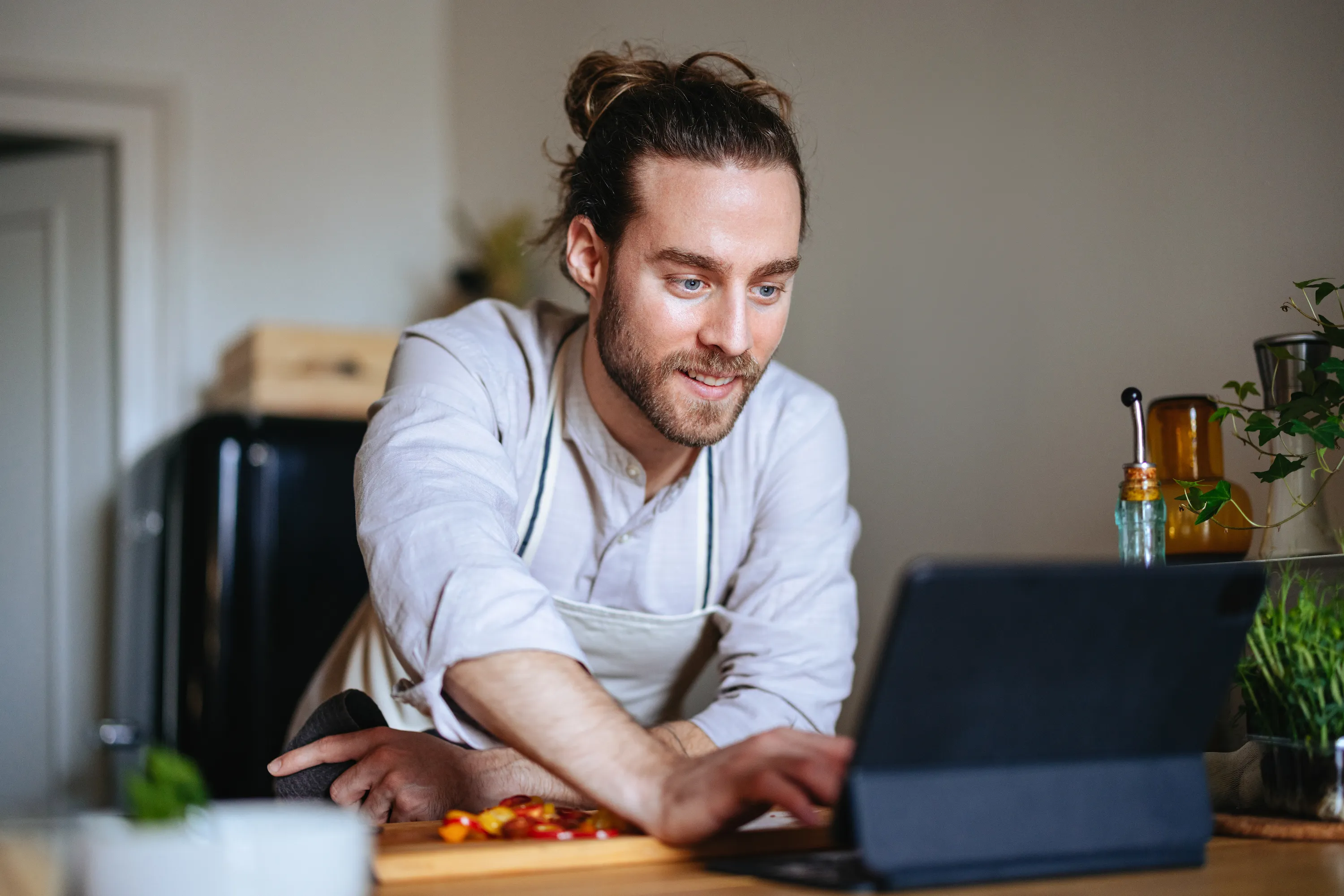 Man in a kitchen wearing an apron, smiling and using a tablet on a wooden table with chopped vegetables.