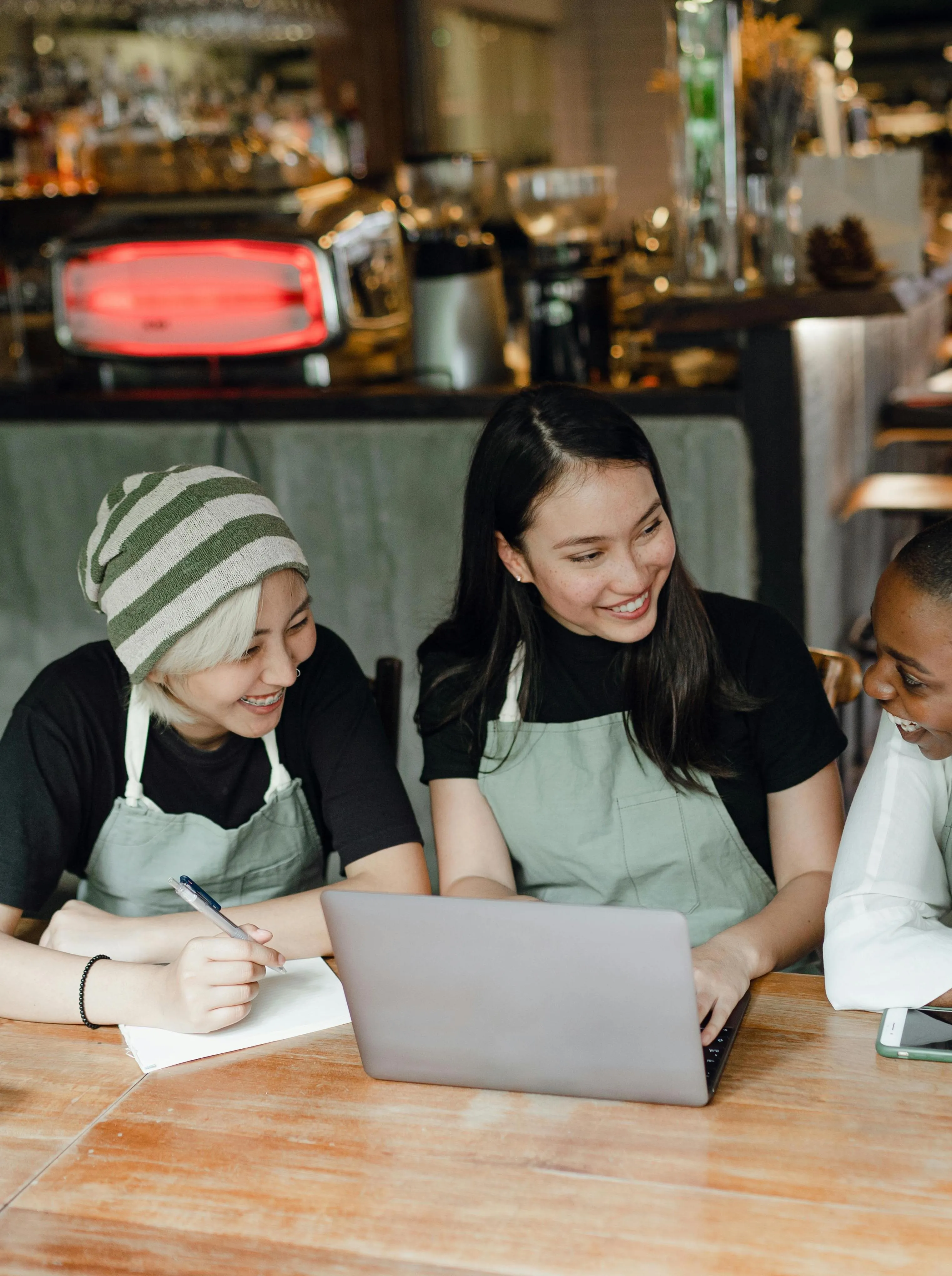 Three women sitting at a wooden table, smiling and working together on a laptop and notebook in a cozy café setting.