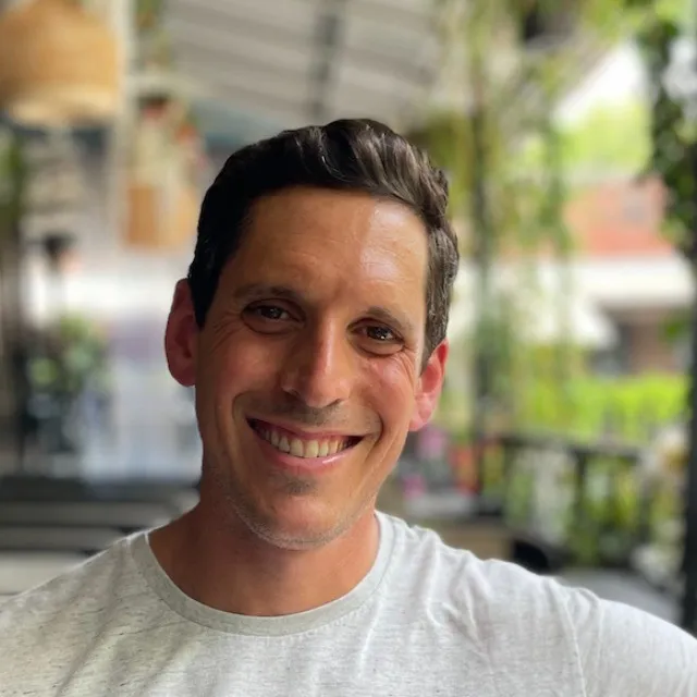 Smiling man with short dark hair wearing a light gray T-shirt, seated outdoors with blurred greenery and structures in the background.