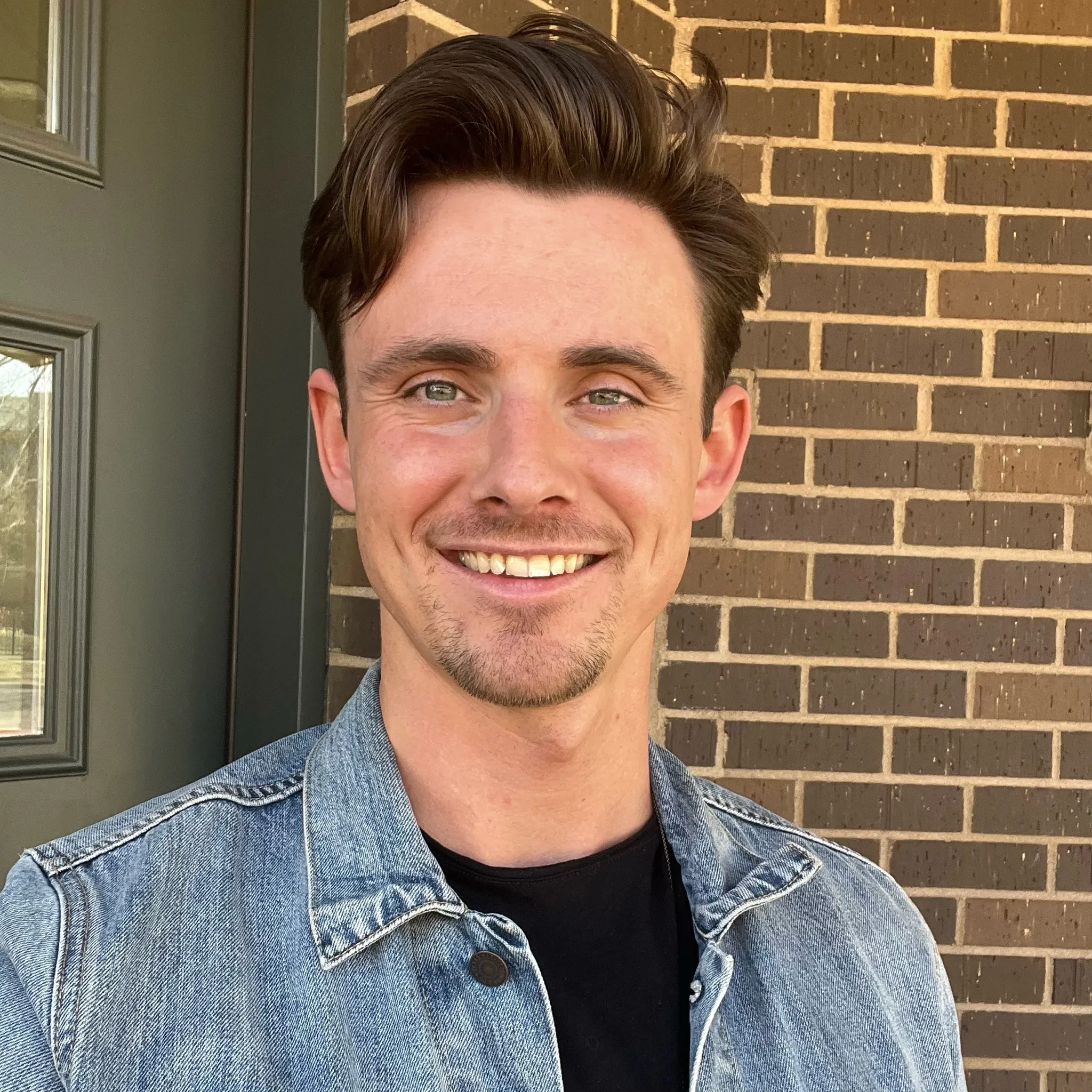Smiling young man with short brown hair, wearing a denim jacket and black shirt, standing in front of a brick wall and a green door.