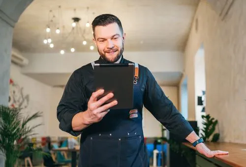 Smiling man in a dark apron looking at a tablet in a modern cafe interior.