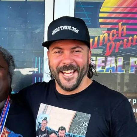 Smiling man with beard wearing a black 'PRO MEAL PREP' cap and a black t-shirt with a photo on it.