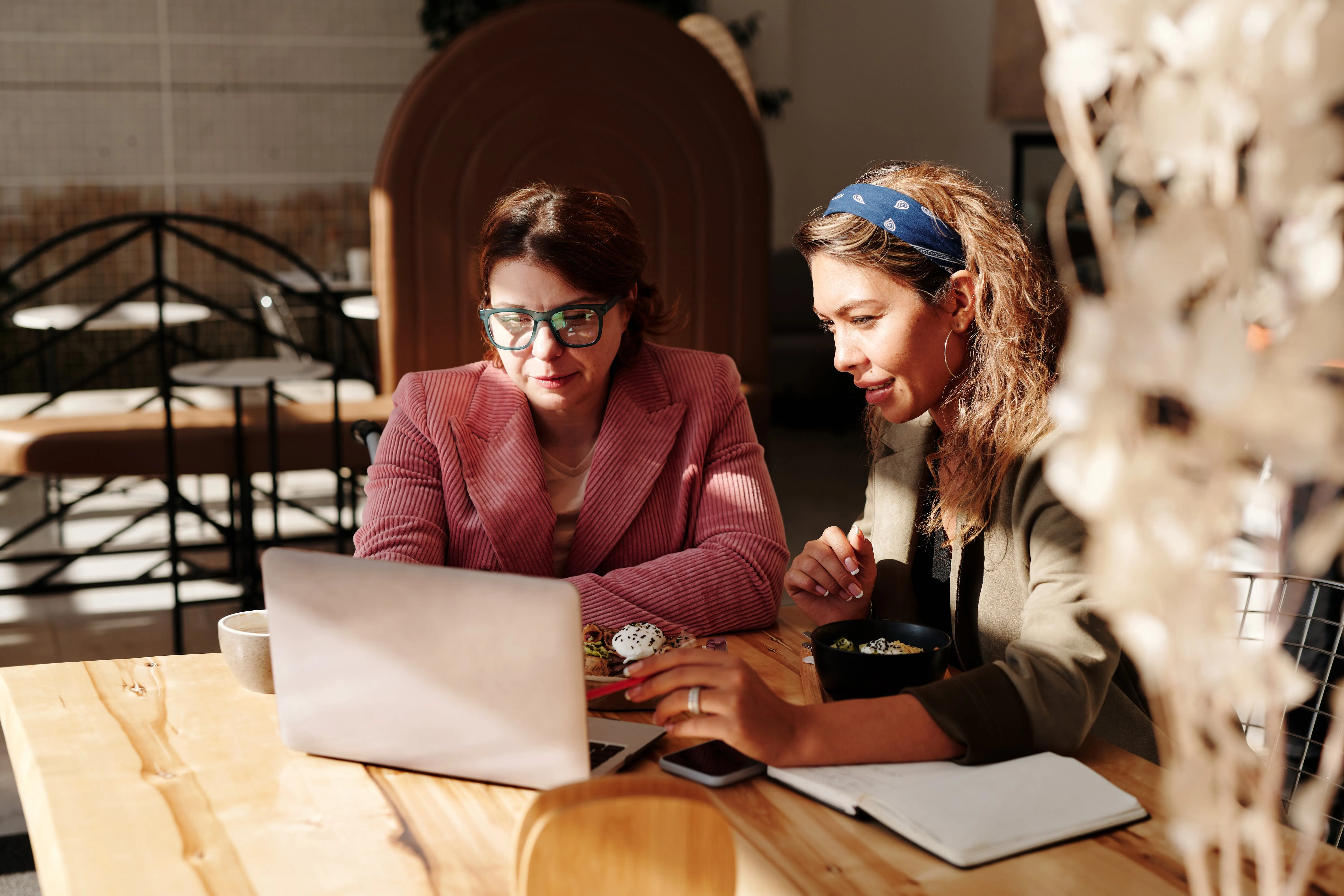 Two women working together on a laptop at a wooden table in a bright cafe.