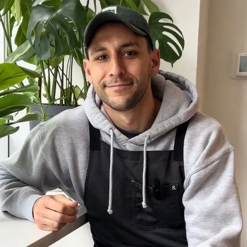 Man wearing a gray hoodie, black apron, and black cap sitting indoors with green plants in the background.