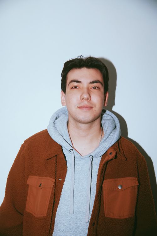 Young man with short dark hair wearing a gray hoodie and a brown jacket against a plain light background.