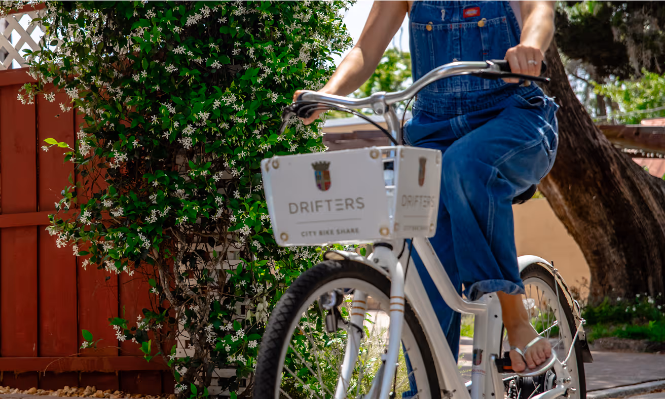 Woman in denim overalls rides a white Drifters City Bike Share bicycle past stone walls and blooming jasmine in St. Augustine.