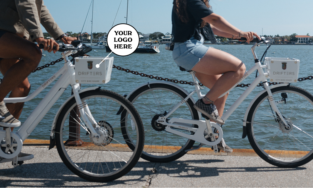 Two cyclists pedal white Drifters City bikes along the Matanzas River, with sailboats and blue sky in the waterfront backdrop.