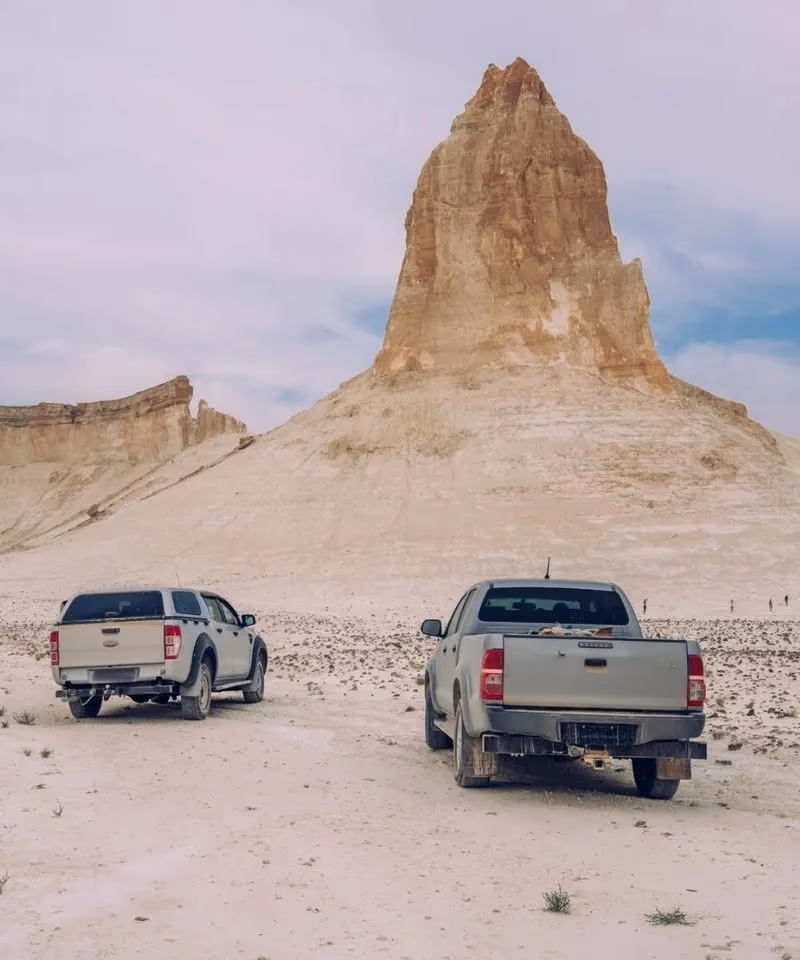 Two off-road pickup trucks parked in front of a giant limestone rock formation at the Boszhira Tract in Mangystau, Kazakhstan.
