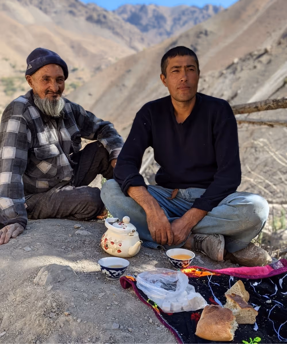 Two local Tajik men sitting outdoors sharing traditional tea and bread in a mountain valley during a group tour.