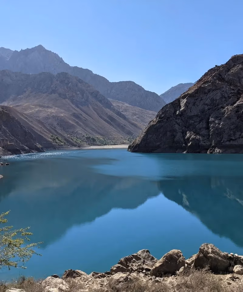 A beautiful turquoise alpine lake surrounded by rugged, dry rocky mountains under a clear blue sky in Tajikistan.