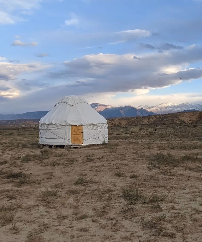 A traditional white nomadic yurt standing on the grassy steppe under a cloudy sky with distant snow-capped mountains.