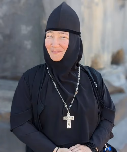 A smiling Orthodox Christian nun in black habit wearing a silver cross necklace in front of a stone monastery wall.