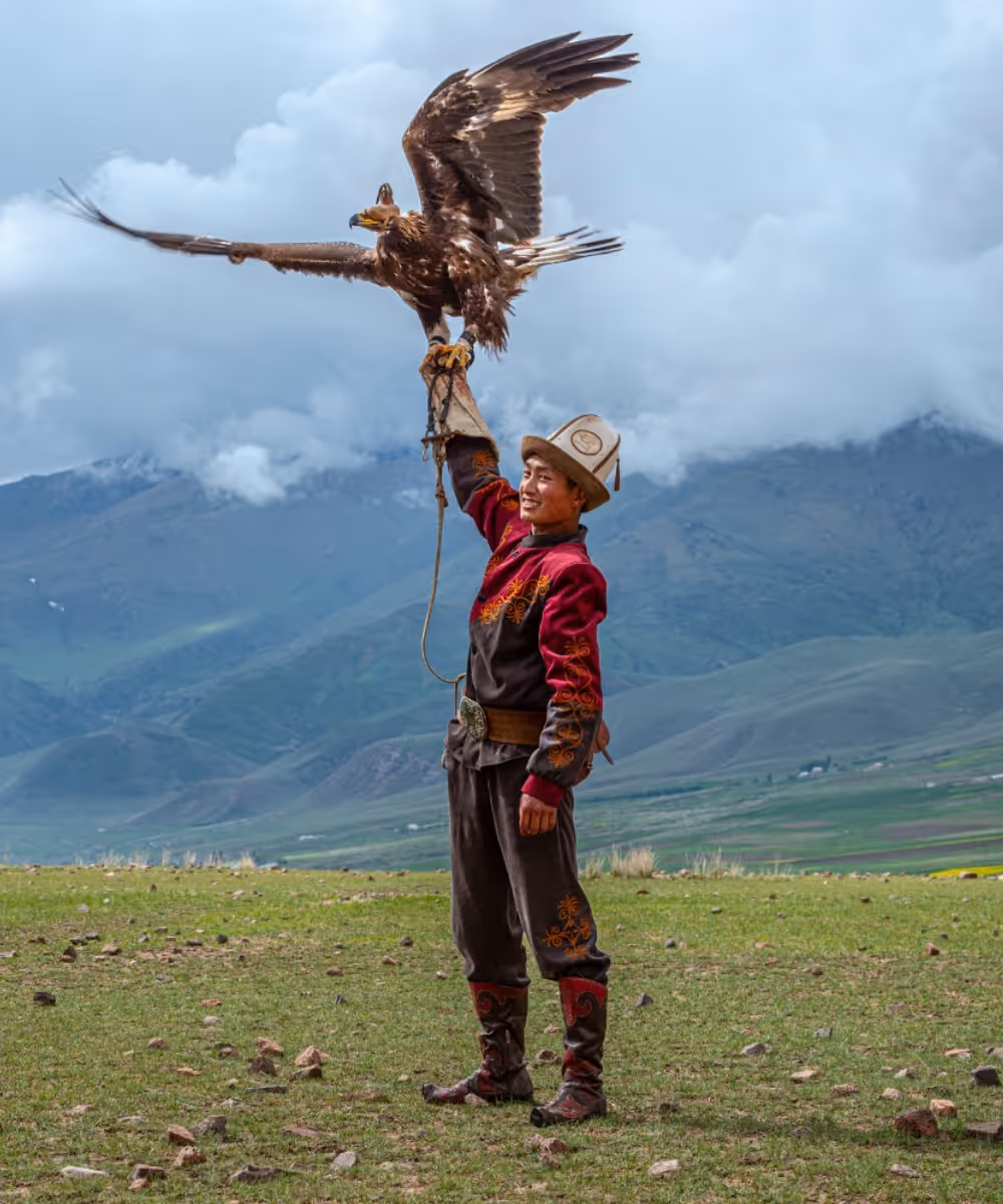 A young Kyrgyz eagle hunter in traditional kalpak hat and embroidered costume holding a large golden eagle with spread wings in Kyrgyzstan.