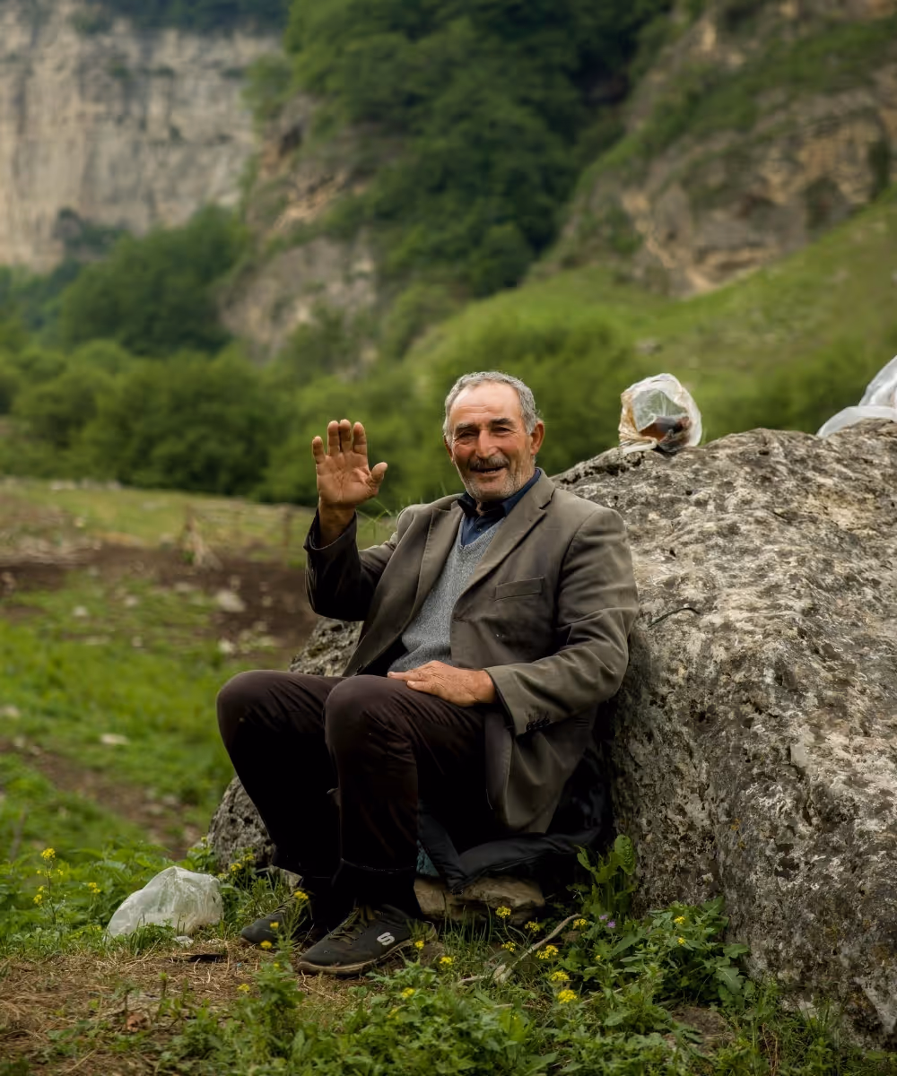 A friendly local man smiling and waving while sitting on a rock in a lush green mountain valley during an Adventure Bound tour.