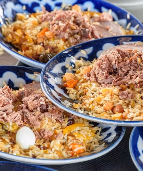 Bowls of traditional Uzbek plov (pilaf rice dish) served with tender meat, carrots, raisins, and quail eggs in blue-patterned ceramic bowls.