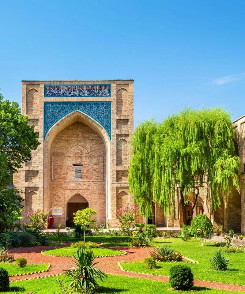 A garden courtyard in front of a blue-tiled ancient madrasah portal in Uzbekistan.
