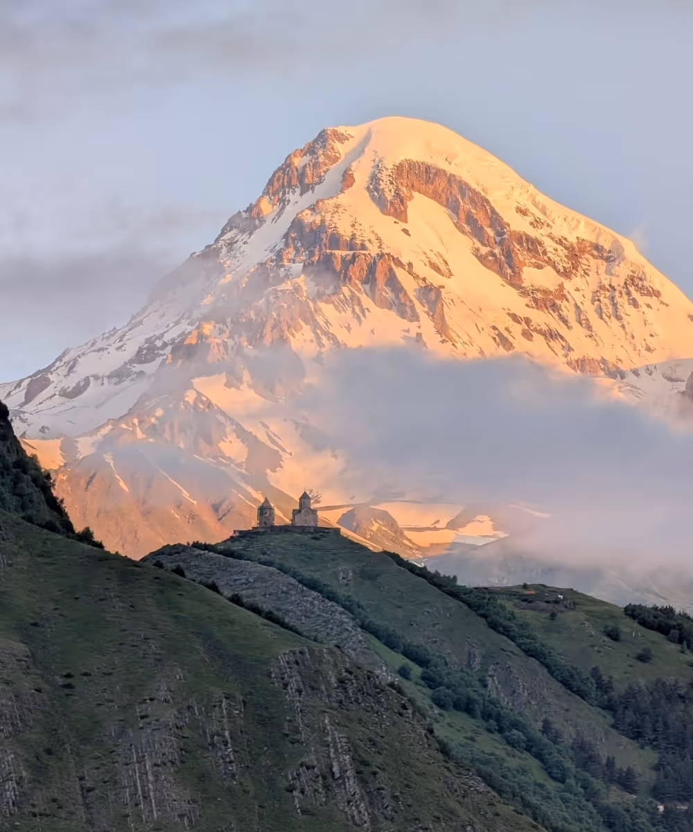 The Gergeti Trinity Church standing on a green hill beneath the snow-capped peak of Mount Kazbek in Georgia.