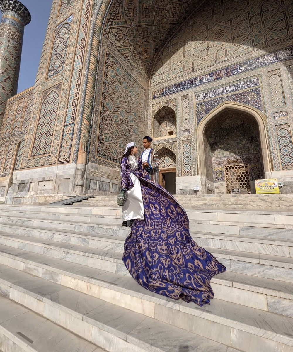 A couple dressed in traditional Uzbek clothing standing on the steps of the Ulugh Beg Madrasah at the Registan in Samarkand, Uzbekistan.