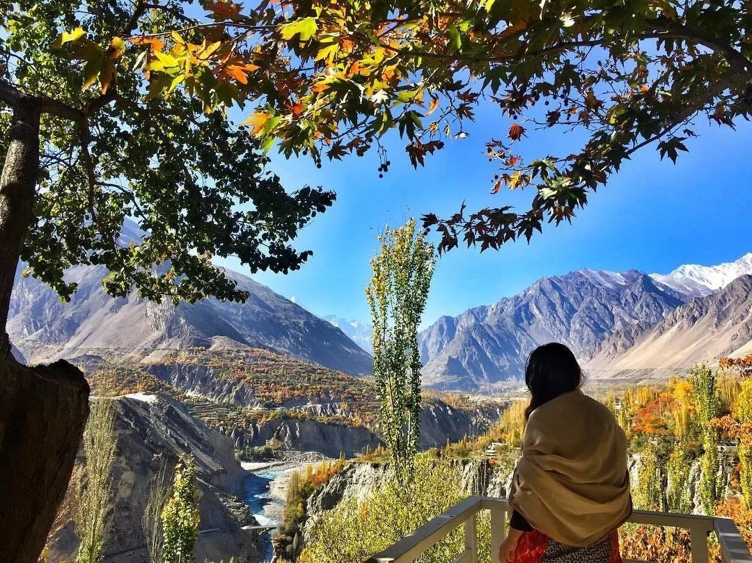 Woman gazes at golden autumn Hunza Valley with vibrant trees and turquoise river below snow peaks.
