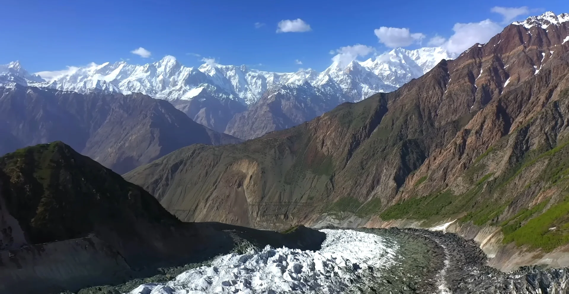 Aerial view of snowy Karakorum Range with a white glacier flowing through deep green valleys.