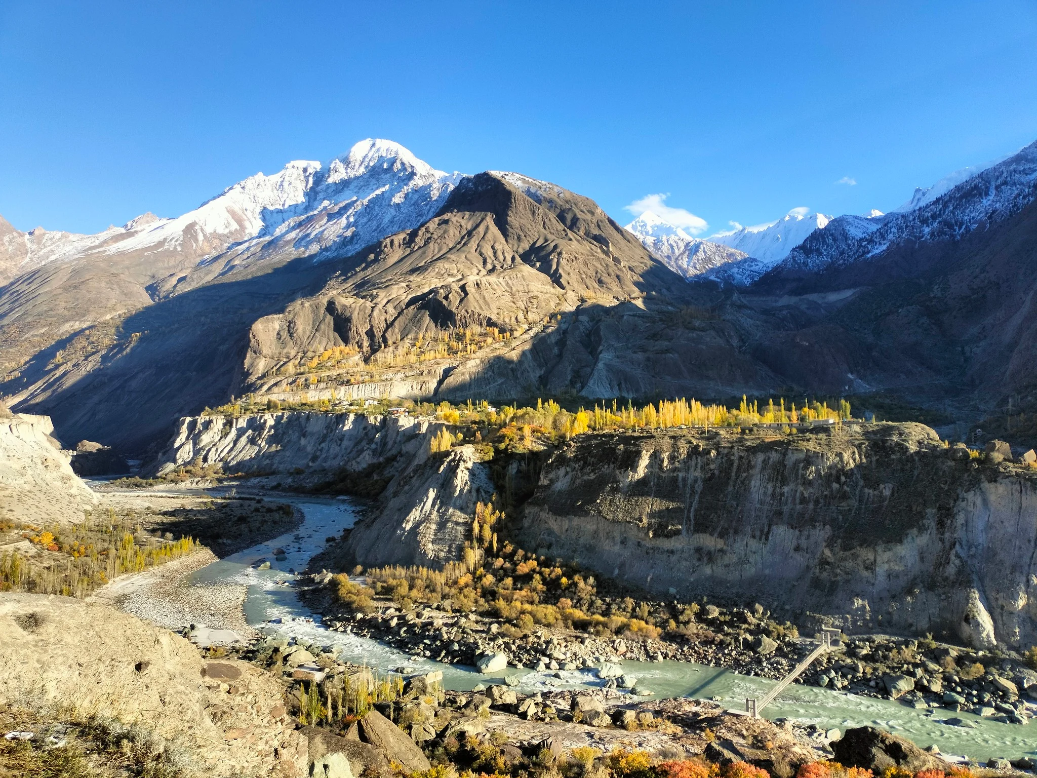 Hunza River winds through a rugged gorge framed by golden autumn trees and snow-capped peaks.