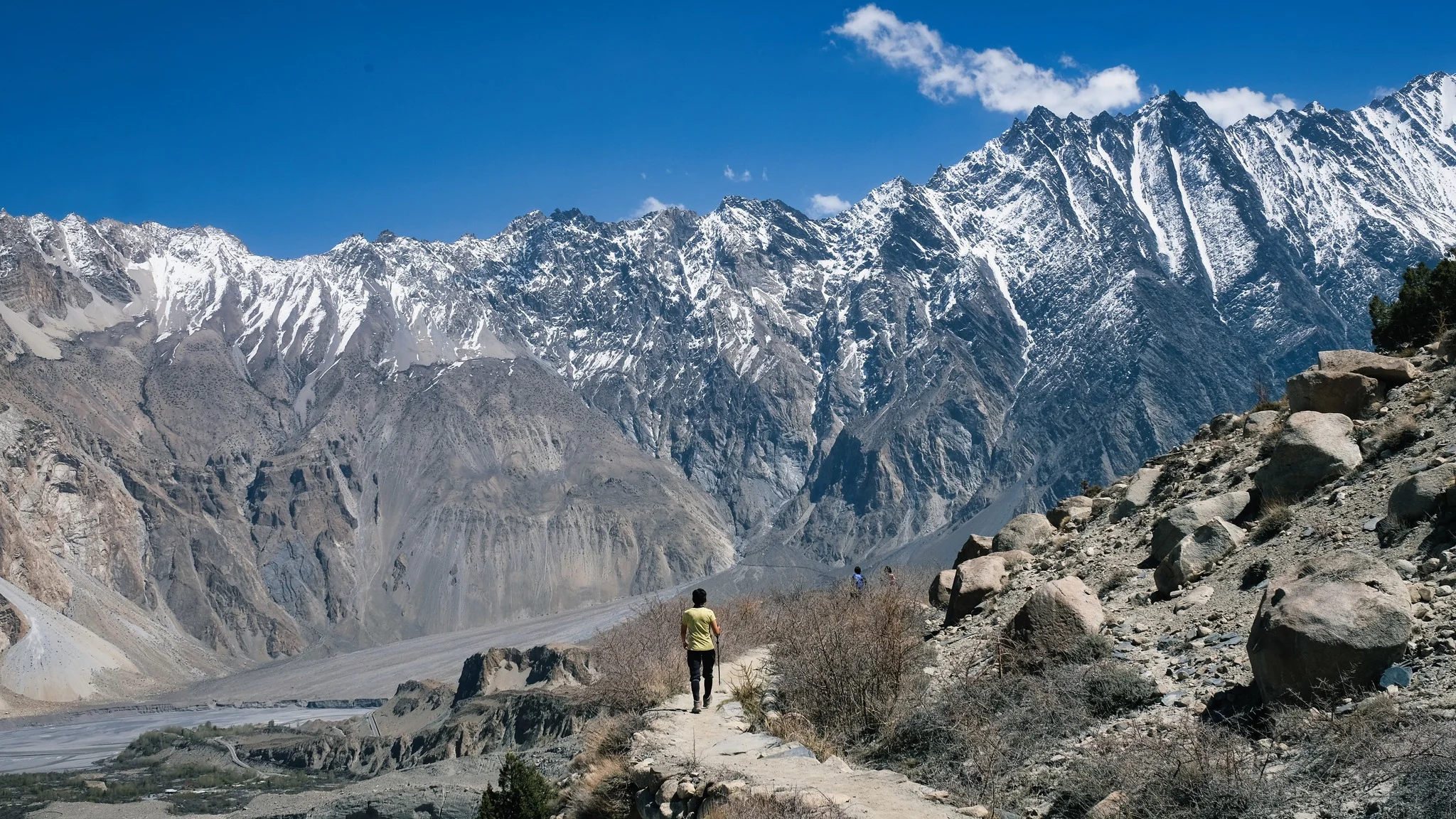 Hiker walks rocky trail with glacier backdrop beneath the towering peaks of Passu Valley.