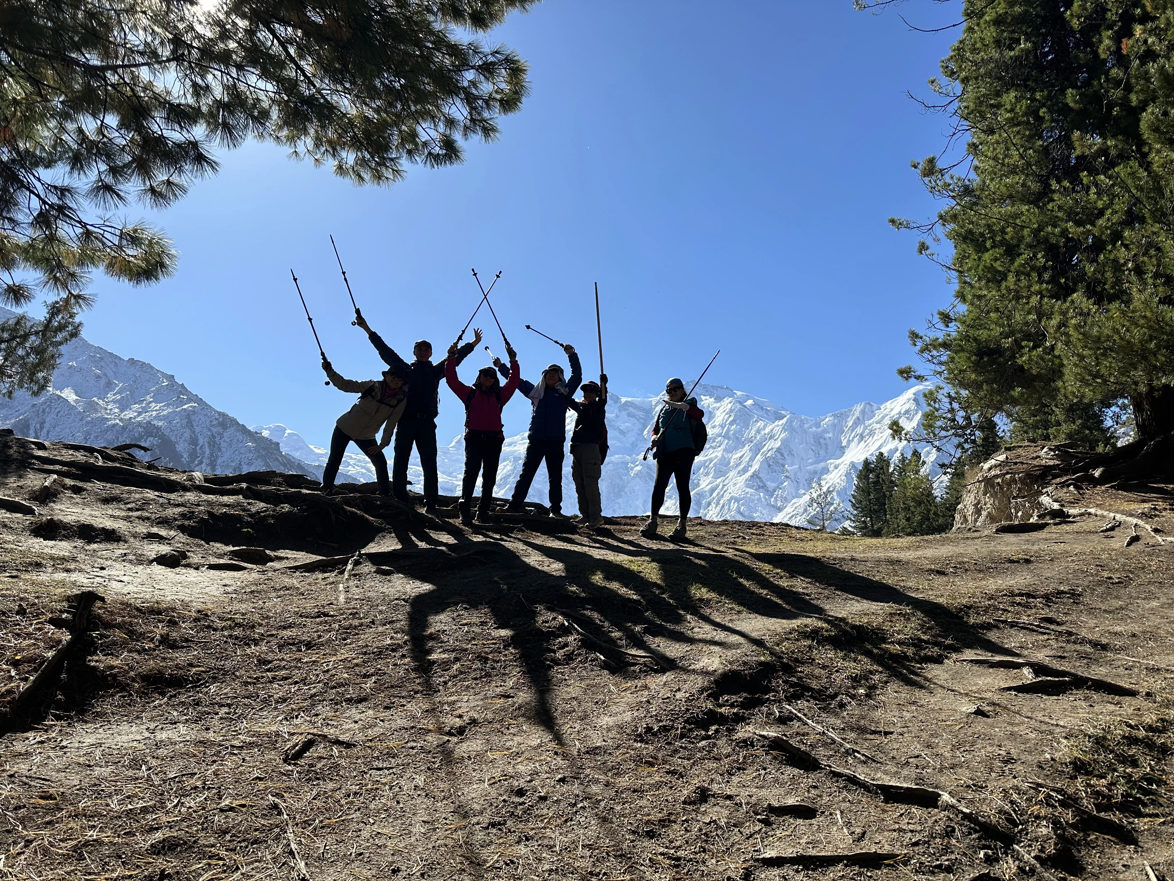 Group of hikers cheer at Nanga Parbat viewpoint with trekking poles raised in triumph.