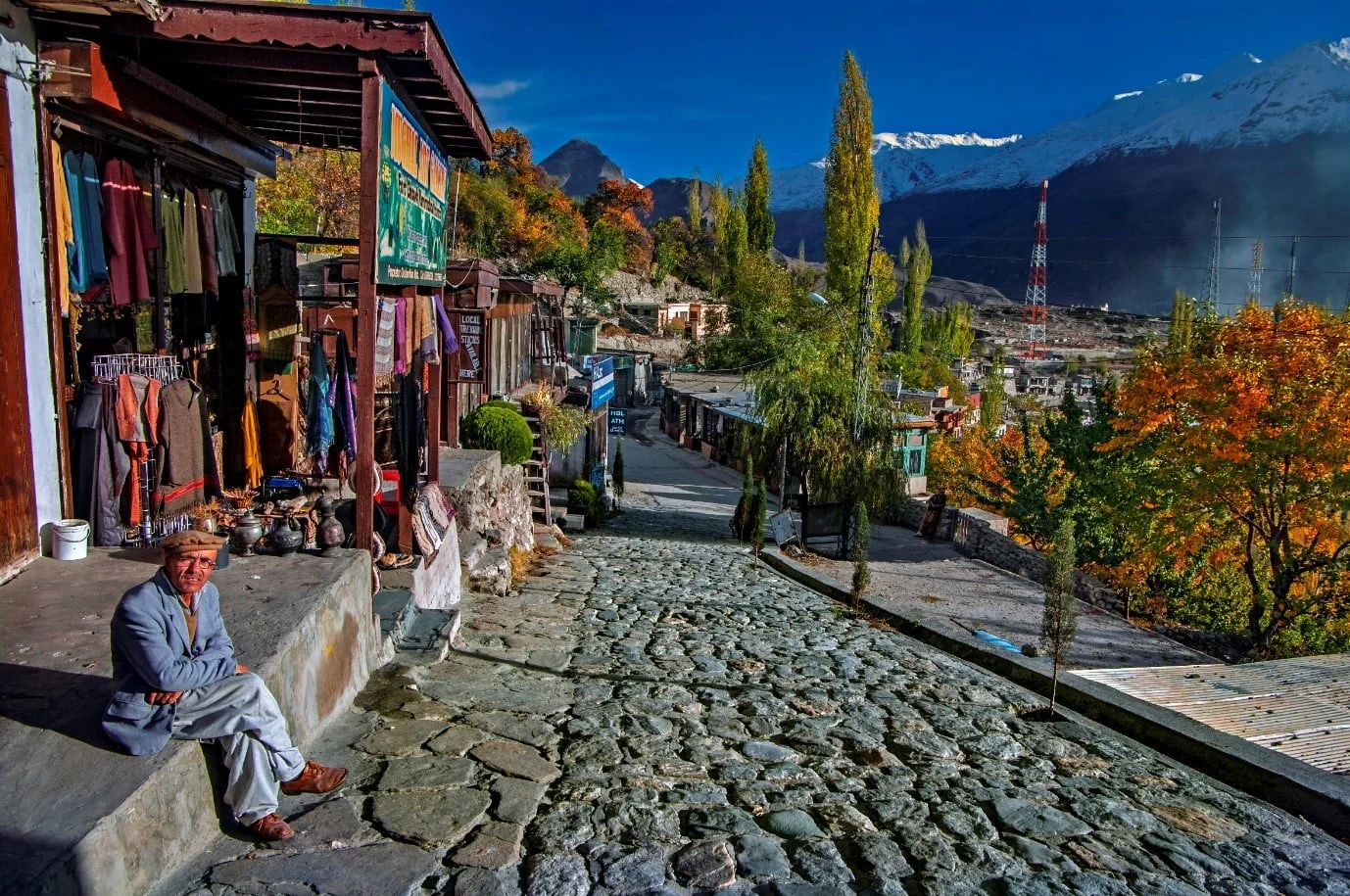 Local shopkeeper sits in Karimabad Bazaar, surrounded by crafts and autumn mountain scenery.